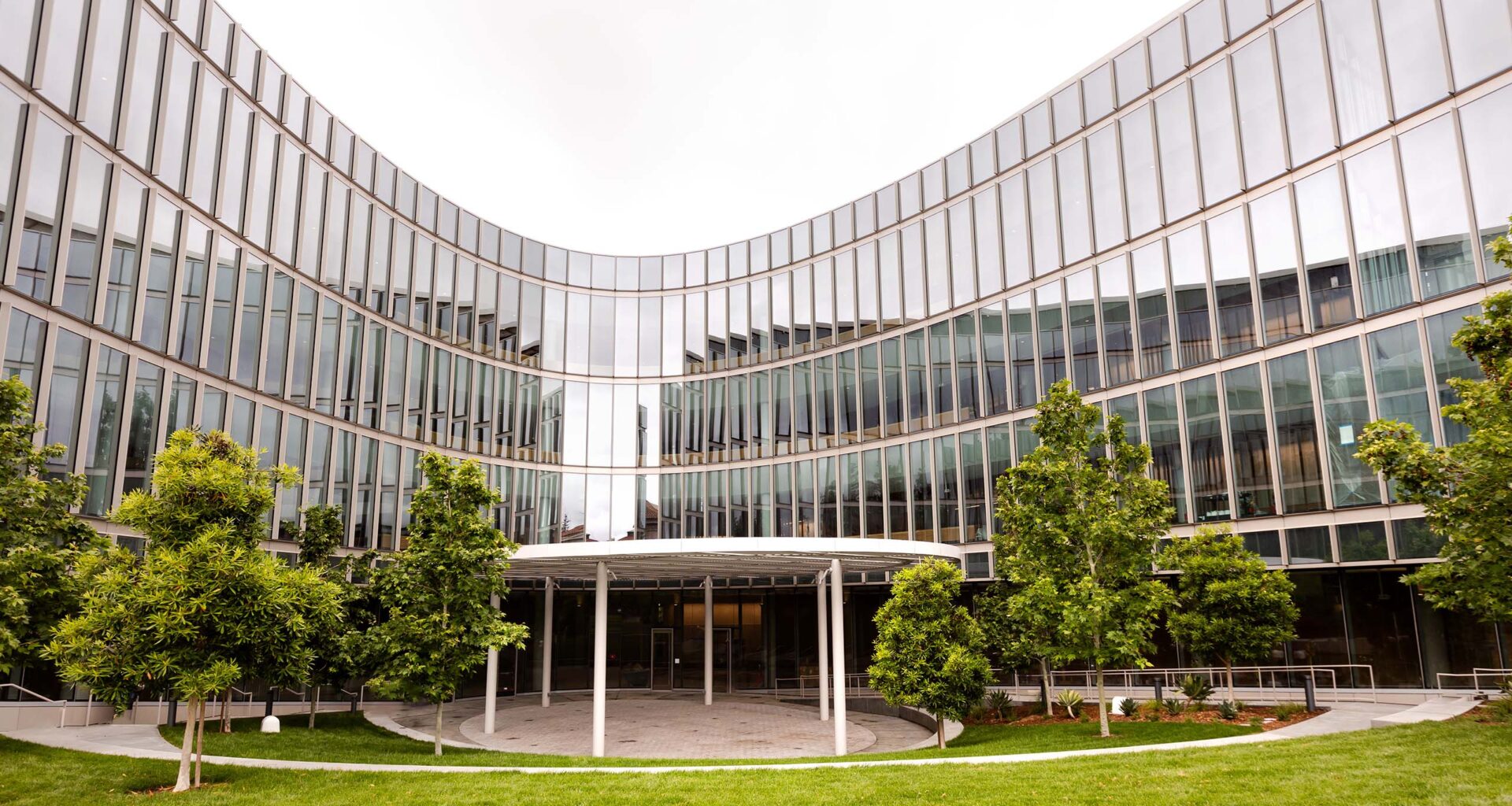 Curved glass exterior and south courtyard of the Barbara and Gerson Bakar Gateway, home of the UC Berkeley College of Computing, Data Science, and Society, featuring a shade canopy, landscaped trees and tiered lawn area.