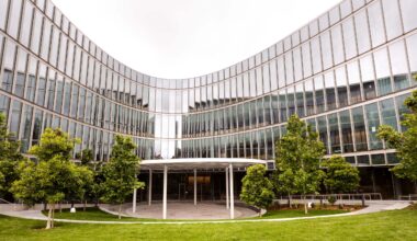 Curved glass exterior and south courtyard of the Barbara and Gerson Bakar Gateway, home of the UC Berkeley College of Computing, Data Science, and Society, featuring a shade canopy, landscaped trees and tiered lawn area.