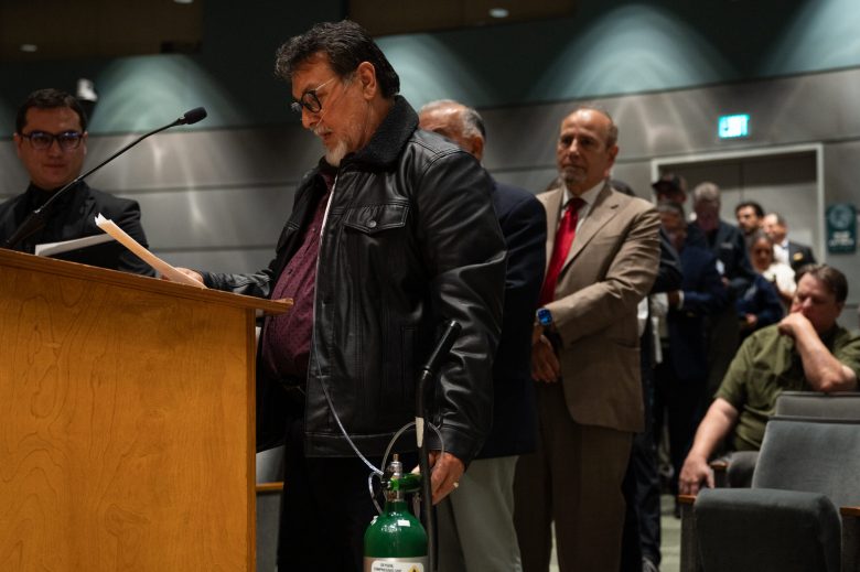 A person in a leather jacket reads at a podium, holding an oxygen tank, with others in suits lined up behind him.