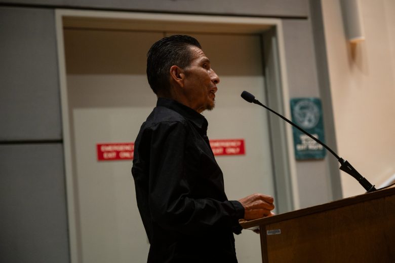 Person speaking at a podium with a microphone, in front of a gray wall and a door with signs.