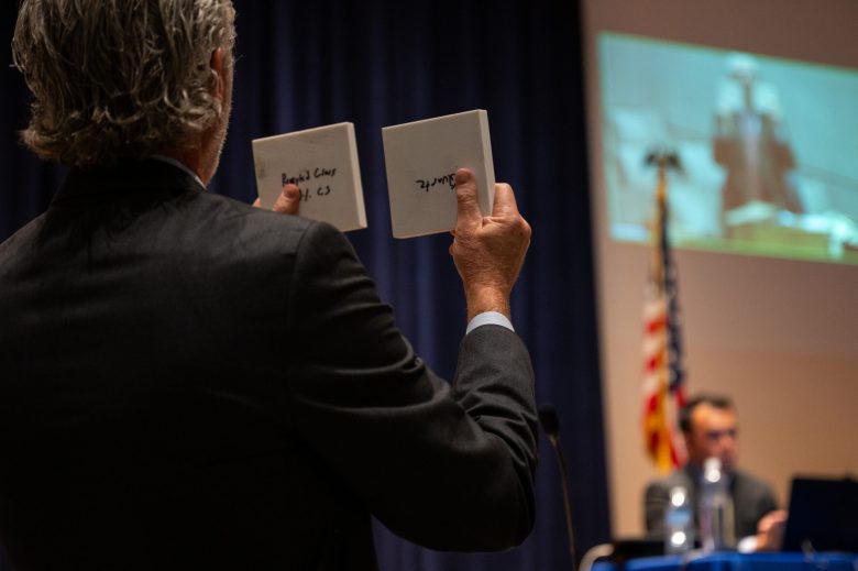 A person in a suit holds up two white plaques with text, blurred audience and a flag in the background.