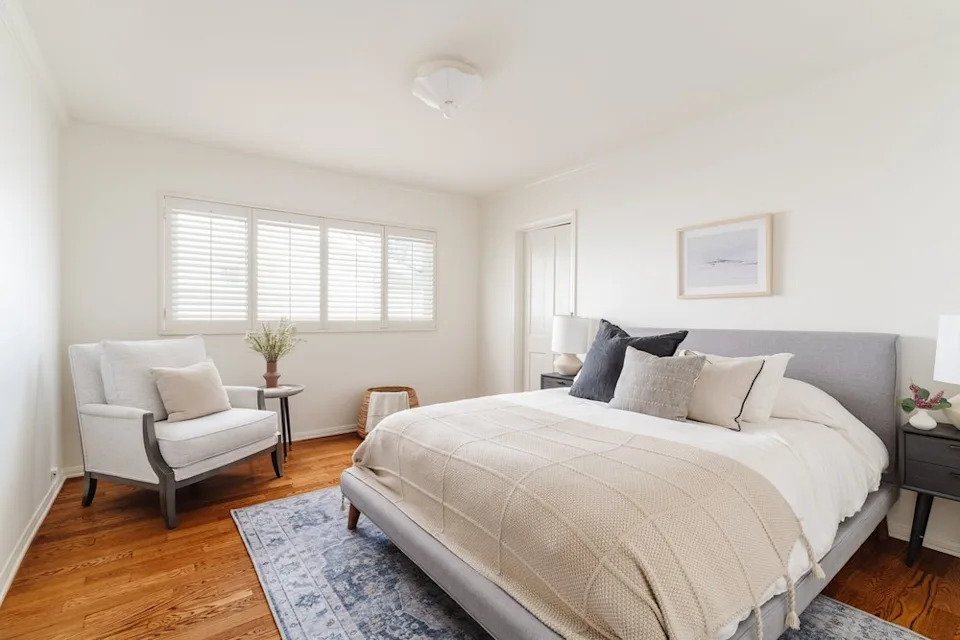 A modern bedroom with a gray fabric bed, a blue patterned area rug, and a white armchair next to a shuttered window.