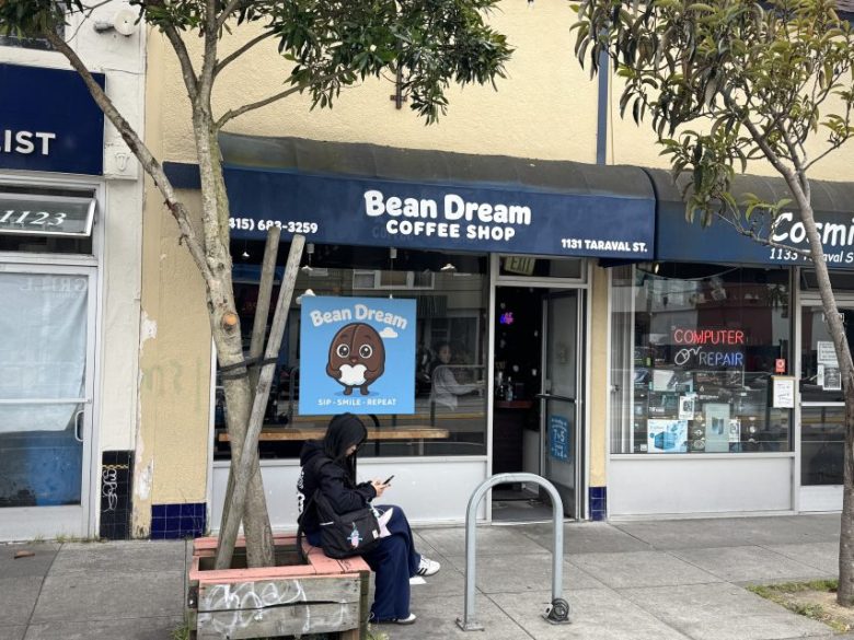 A person sits on a bench using a phone outside Bean Dream Coffee Shop, which has a blue sign and awning on a city street.