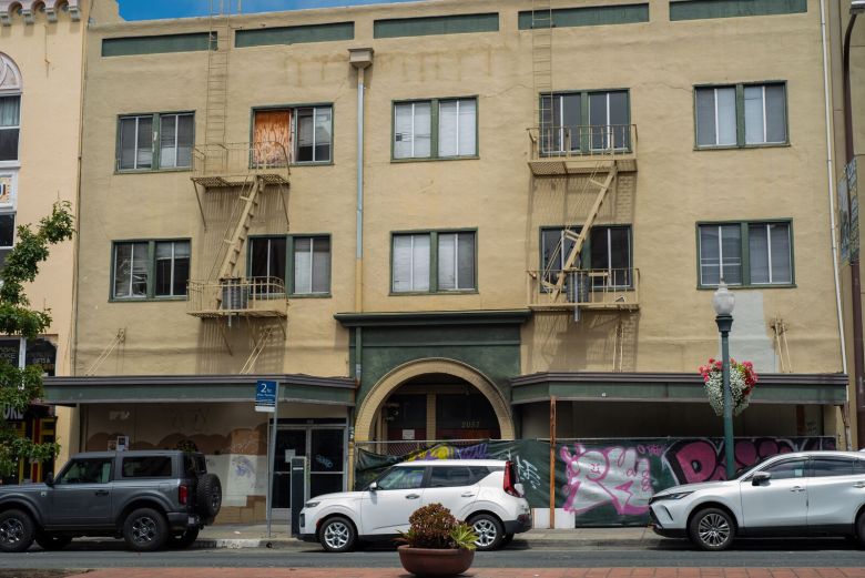 A view of a three-story apartment building. One of the building's windows is boarded up, and its ground floor is blocked by a graffiti-covered fence