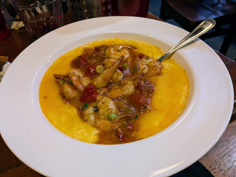 A white bowl filled with creamy yellow grits topped with shrimp in a brown sauce, garnished with green onions and diced tomatoes, with a spoon on the side.
