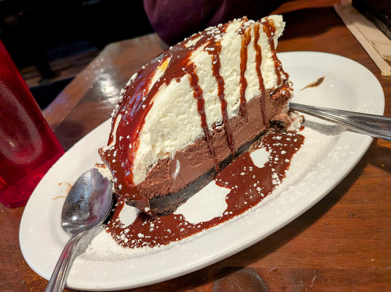A slice of layered chocolate and cream pie with chocolate sauce and powdered sugar on a white plate, with two spoons beside it.