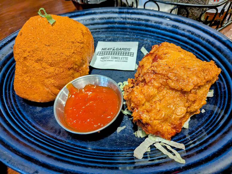 A piece of fried chicken and a bread roll coated in seasoning on a blue plate, served with a cup of red sauce, shredded lettuce, and a moist towelette.
