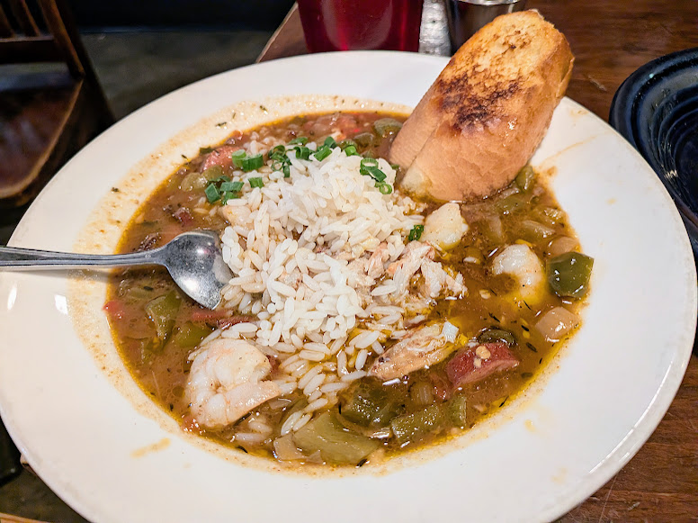 A bowl of gumbo with shrimp, rice, vegetables, and green onions, served with a slice of toasted bread on a white plate.