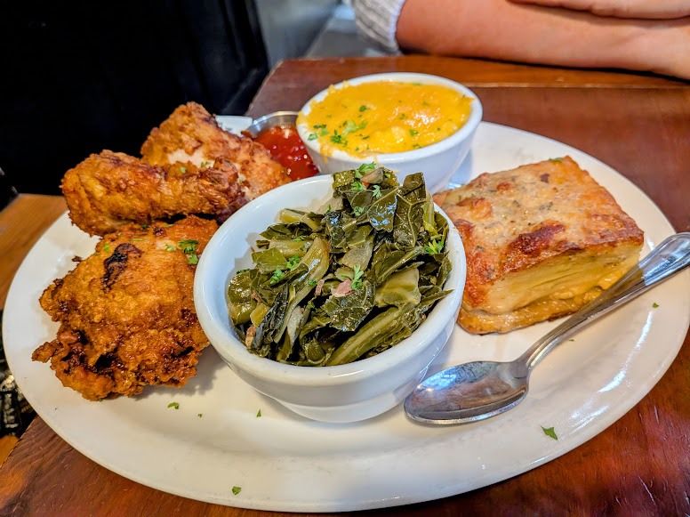 A plate with fried chicken, collard greens, macaroni and cheese, and a slice of potato gratin, served on a wooden table with a spoon.