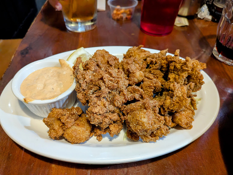A plate of fried oysters served with a side of dipping sauce on a white dish, set on a wooden table with drinks in the background.