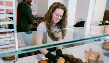 A smiling woman with curly hair and glasses wears a gray shirt and dark apron, standing with crossed arms inside a colorful room with food on a table.