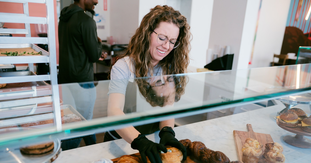A smiling woman with curly hair and glasses wears a gray shirt and dark apron, standing with crossed arms inside a colorful room with food on a table.