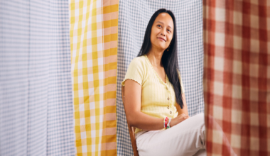 A woman with long black hair sits cross-legged on a wooden chair, wearing a yellow top, white pants, and silver ballet flats against a backdrop of checkered fabrics.