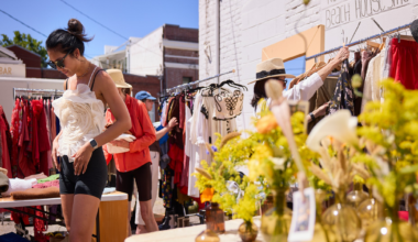 A man and a woman examine items on a wooden table filled with various antiques and household goods at an outdoor market.