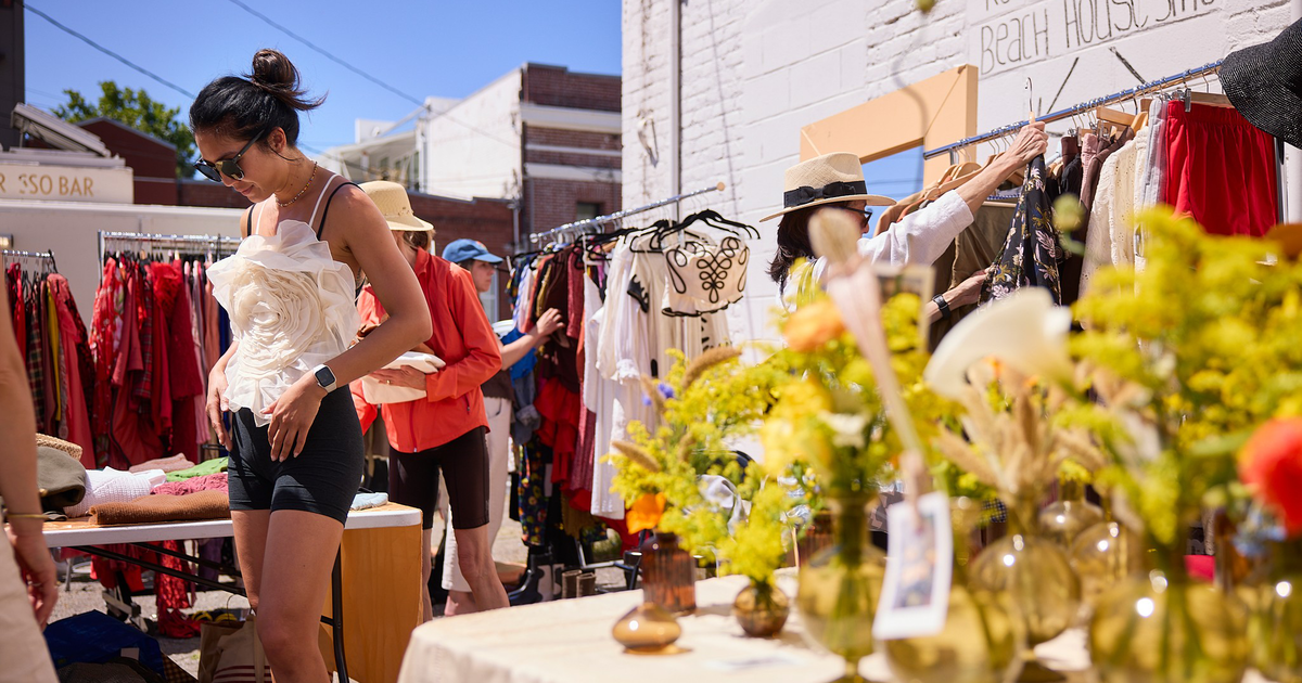 A man and a woman examine items on a wooden table filled with various antiques and household goods at an outdoor market.