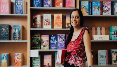 A smiling woman with long dark hair wears a red and white polka dot top, standing in front of wooden shelves filled with various colorful books.