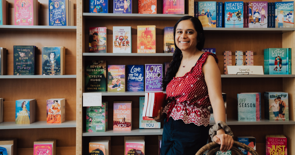 A smiling woman with long dark hair wears a red and white polka dot top, standing in front of wooden shelves filled with various colorful books.