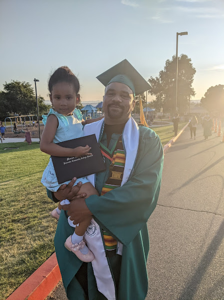 Long wearing graduation regalia during festivities for the Moreno Valley College. He's holding his daughter, who is holding his diploma holder.