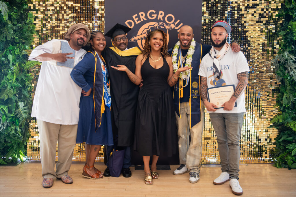 A group of six people stand in front of a sign that says "Underground Scholars." They're celebrating a recent graduation.