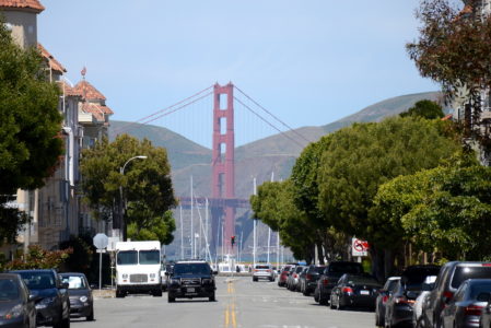 A view of the Golden Gate Bridge seen from a city street lined with trees, parked cars, and residential buildings on a clear day.