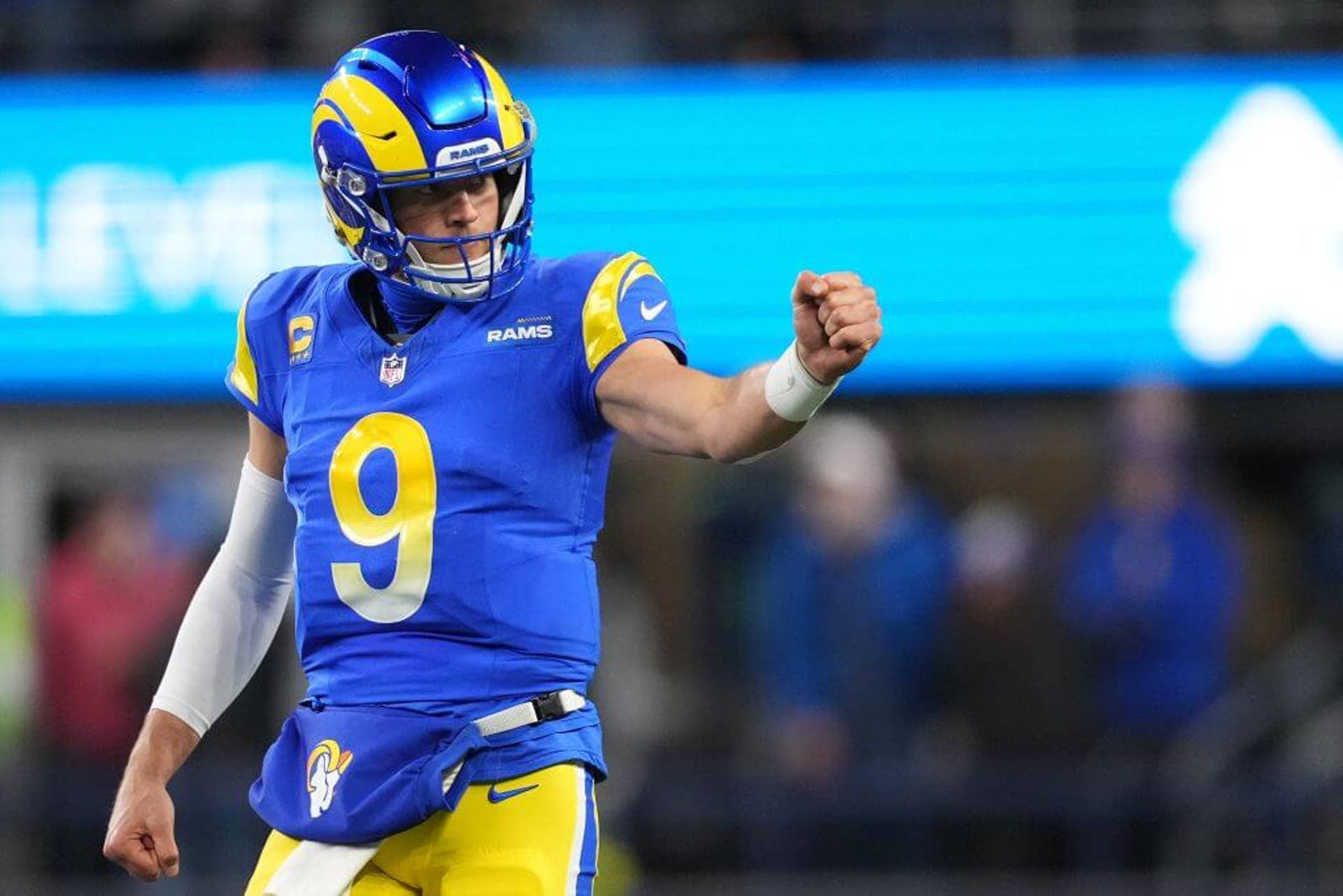 Matthew Stafford, wearing a blue No. 9 Rams uniform, looks to his left while holding out a clenched left fist.