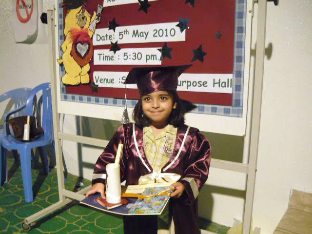 A young Hannah Rabbani at her primary school graduation in 2010. She is wearing a maroon and white cap and gown, holding a diploma and a tray of gifts while smiling proudly in front of a colorful school bulletin board.