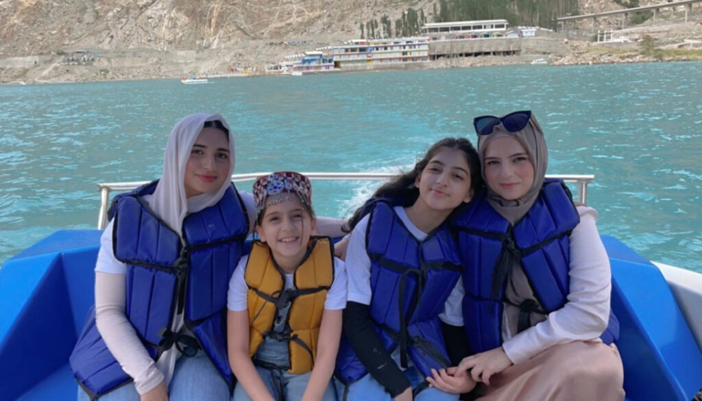 Hannah Rabbani (left) with her two younger sisters and mom (right) on a boat. They are all wearing life jackets, smiling against a backdrop of a clear turquoise lake and rugged, towering mountains.