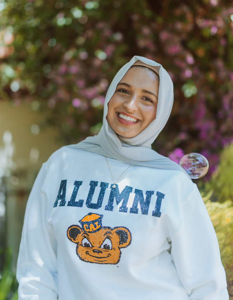 Hannah Rabbani wearing a Cal Berkeley alumni sweatshirt and a light grey hijab, smiling in front of a background of flowering trees.