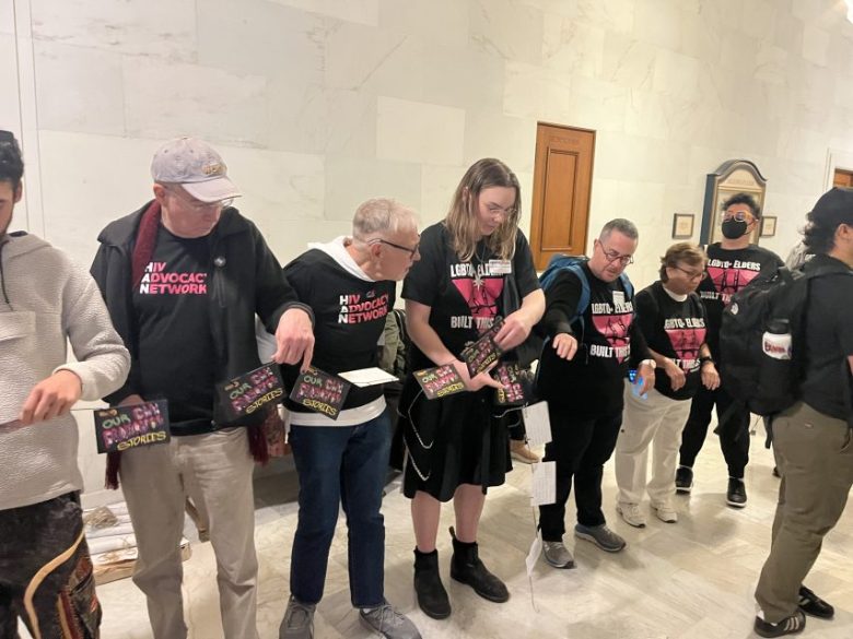 A group of people wearing matching advocacy shirts stand in a hallway, holding small boxes labeled "Shouts" and "Own Your Strength.