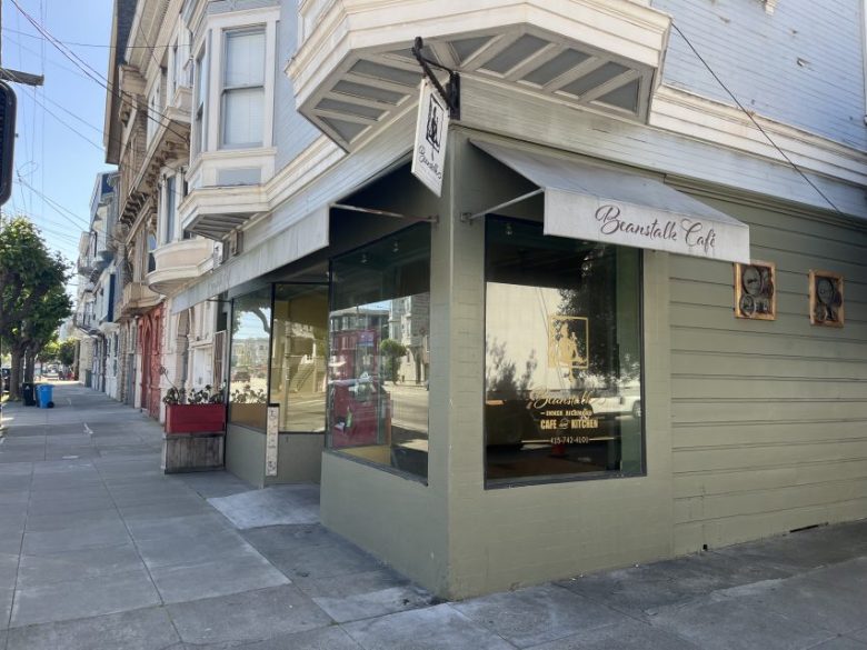 Street view of the exterior of Beansville Café, with large windows, a hanging sign, and a beige awning on the corner of a city block.