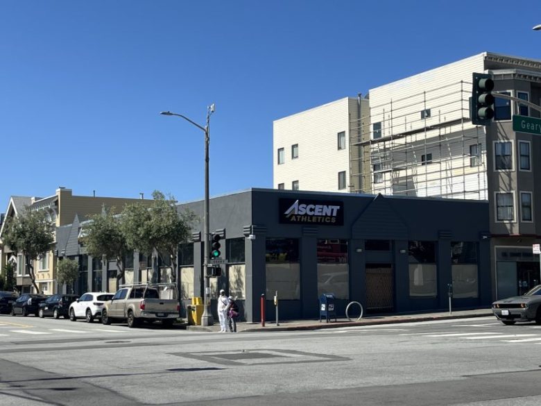 Street view of a corner building with an "Ascent Athletics" sign, parked cars, a traffic light, pedestrians, and scaffolding on an adjacent building under clear blue sky.
