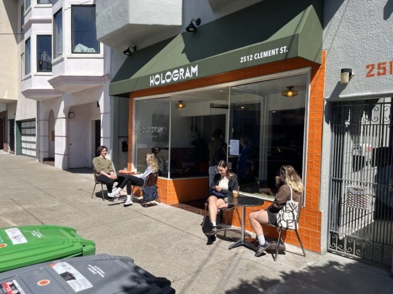 People sit at outdoor tables in front of Hologram café on a sunny day at 2512 Clement St. Some are talking, while others use their phones. The café has an olive green awning and orange tiles.