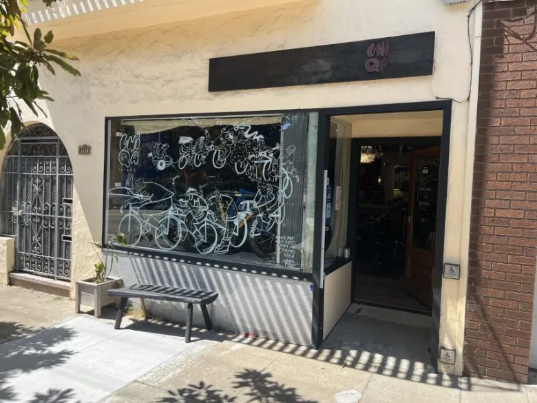 A storefront with a black sign, large window featuring abstract white designs, a black bench outside, and an open door leading inside.