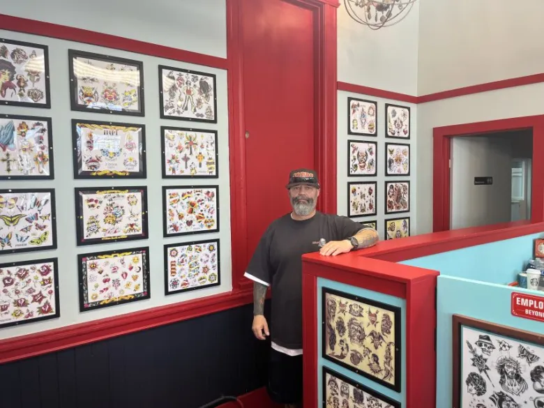 A man stands inside a tattoo shop next to a red counter, with framed tattoo designs covering the walls behind him.