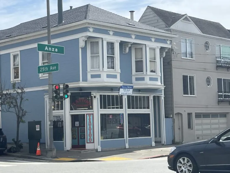 A blue two-story building on a street corner houses a tattoo parlor, with signs for Anza Street and 25th Avenue visible. A traffic light and parked car are in the foreground.