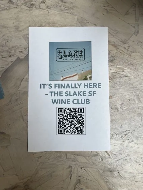 A flyer on a wooden surface announces the launch of the Slake SF Wine Club, featuring a logo, a QR code, and an image of a street scene.