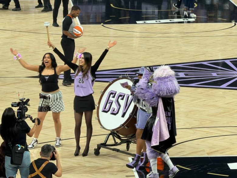 Two women and a costumed mascot stand near a large drum on a basketball court, with one woman raising her arms and a camera crew filming.