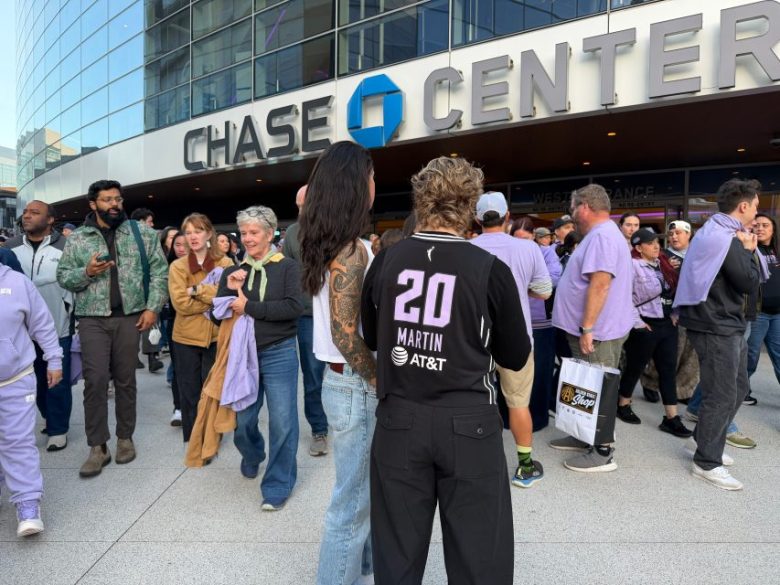 A crowd of people gathers outside the Chase Center; one person in the foreground wears a black "Martin 20" AT&T jersey.