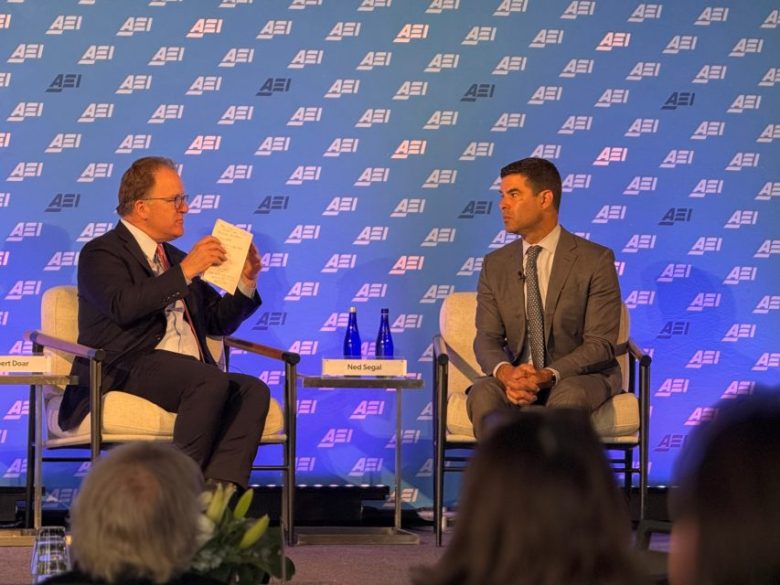 Two men in business attire sit onstage in conversation at an AEI event, with water bottles on a table and an AEI-branded backdrop behind them.