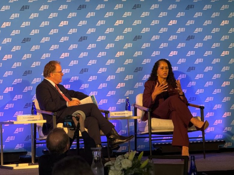 Two people sit on stage in chairs having a discussion at an event with an AEI-branded backdrop. One person gestures while speaking; audience members watch and take photos.