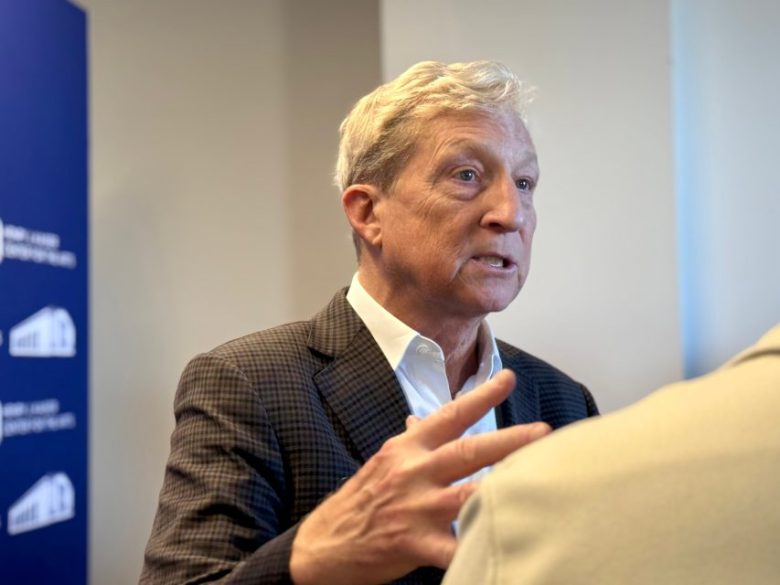 An older man in a checked suit jacket gestures with his hand while speaking to another person indoors.