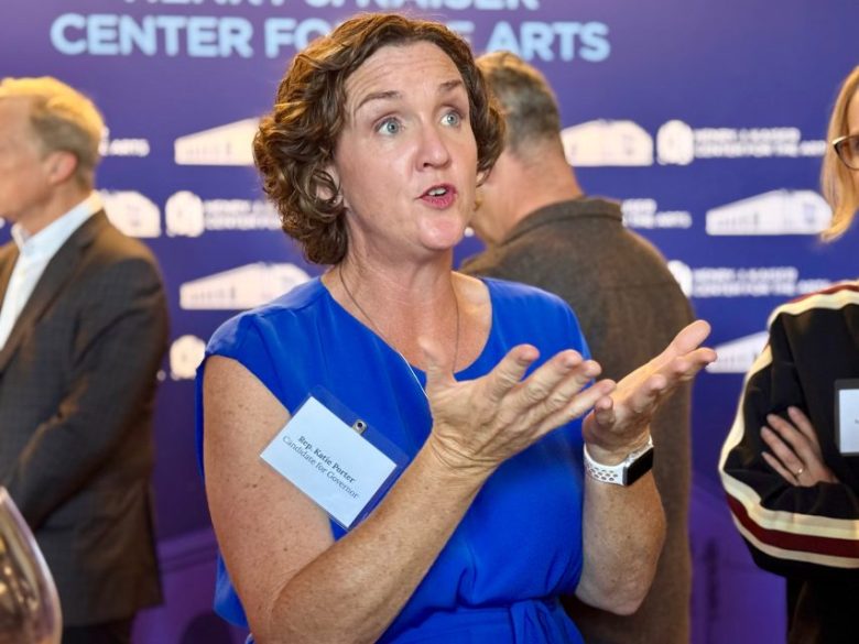 A woman in a blue dress speaks and gestures with her hands at an indoor event, wearing a name badge. Other people are in the background.