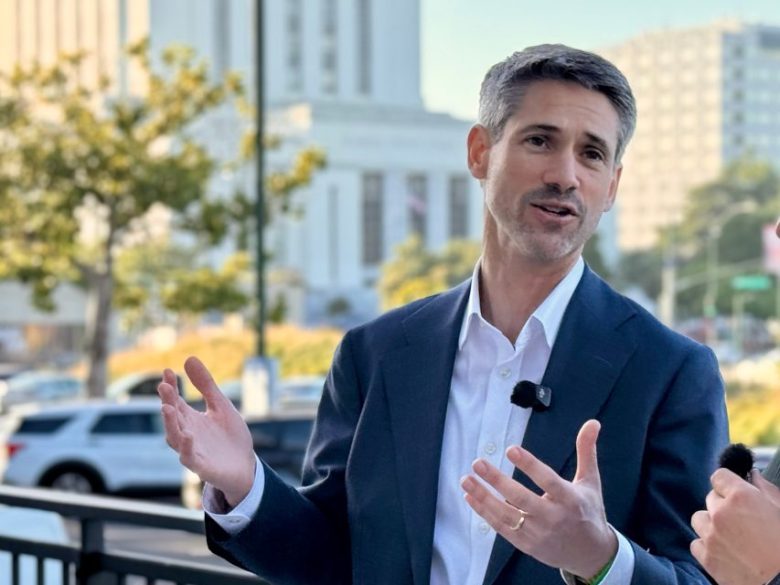 A man in a suit speaks outdoors, gesturing with his hands. He wears a microphone on his lapel. Cars and buildings are visible in the background.