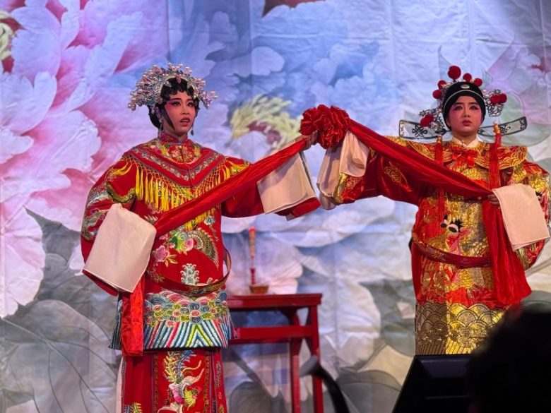 Two performers in traditional Chinese opera costumes hold a red ribbon on stage with a floral backdrop behind them.