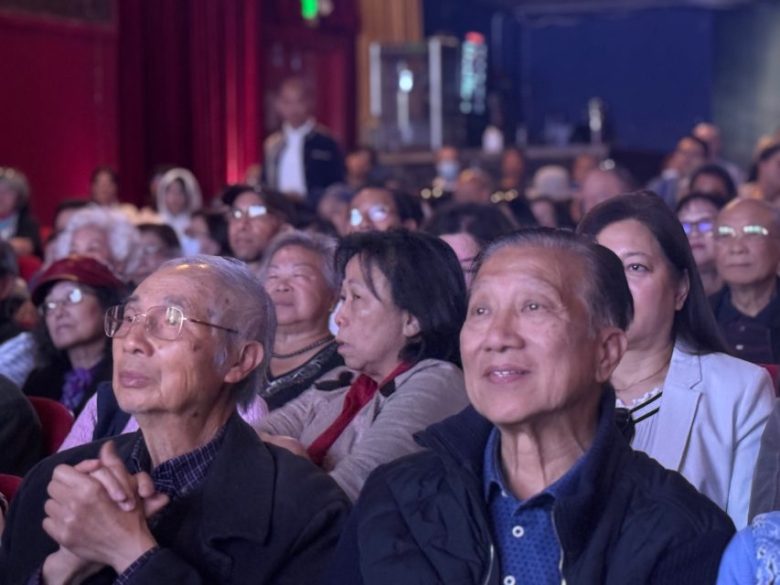 A group of people, mostly older adults, sit closely together in an auditorium, attentively watching something off camera.