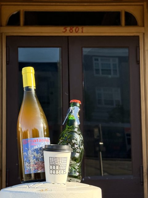 A bottle of Matassa white wine, a green glass bottle shaped like a figure, and a disposable cup labeled "NRG" on a table in front of a building entrance.