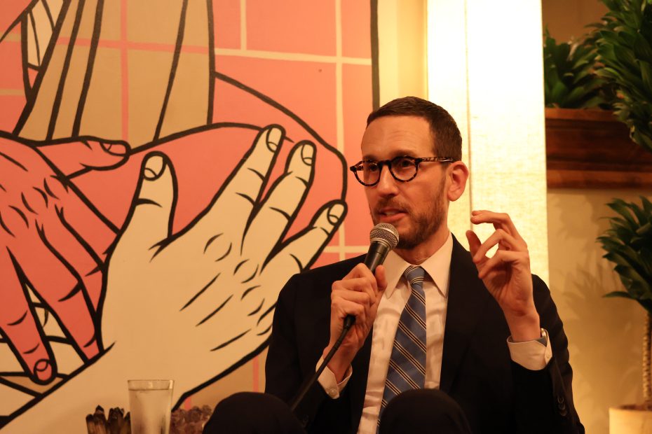 A person in glasses and a suit holds a microphone, speaking in front of a colorful background with large artwork of hands.