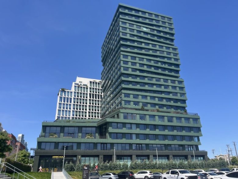 A modern, multi-story green office building with staggered balconies and large windows, set against a clear blue sky with parked cars in the foreground.
