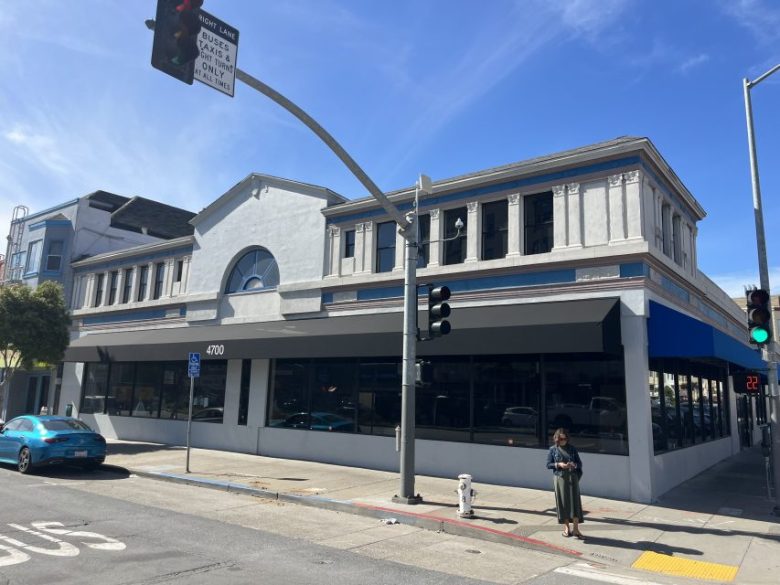 A large, two-story, gray building with blue trim on a street corner, featuring large windows, a traffic light, and a person standing on the sidewalk.
