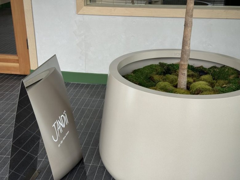 A large white planter with a tree and moss, next to a silver signboard displaying the text "JANDI" in a modern indoor setting.